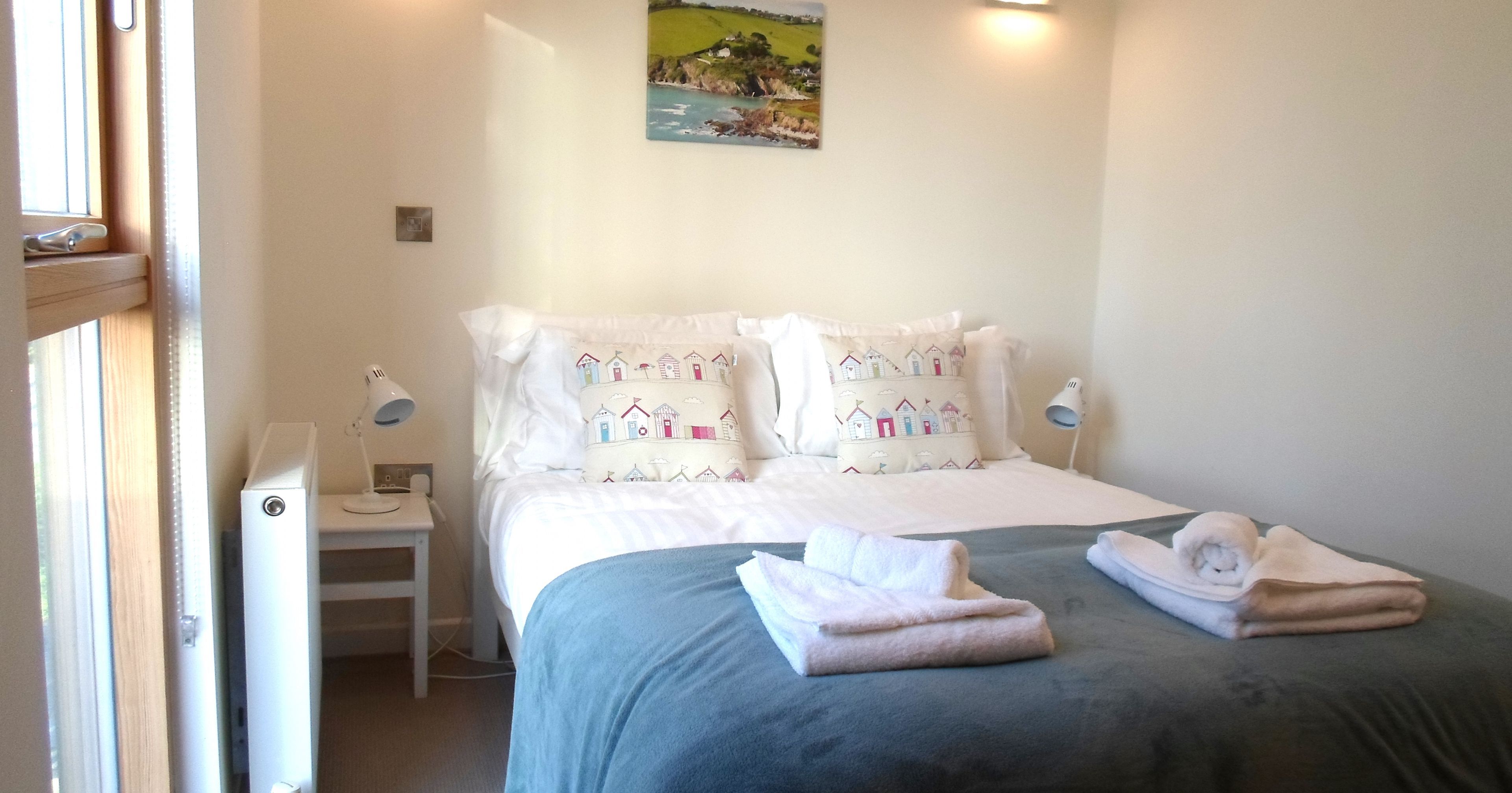 Bright bedroom with double bed, white bedding, colorful pillows, folded towels, and bedside lamps