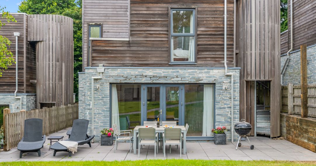 Modern two-story house with wood and stone exterior, glass patio doors, outdoor dining table, lounge chairs, and barbecue grill on a tiled patio, surrounded by grass and plants.