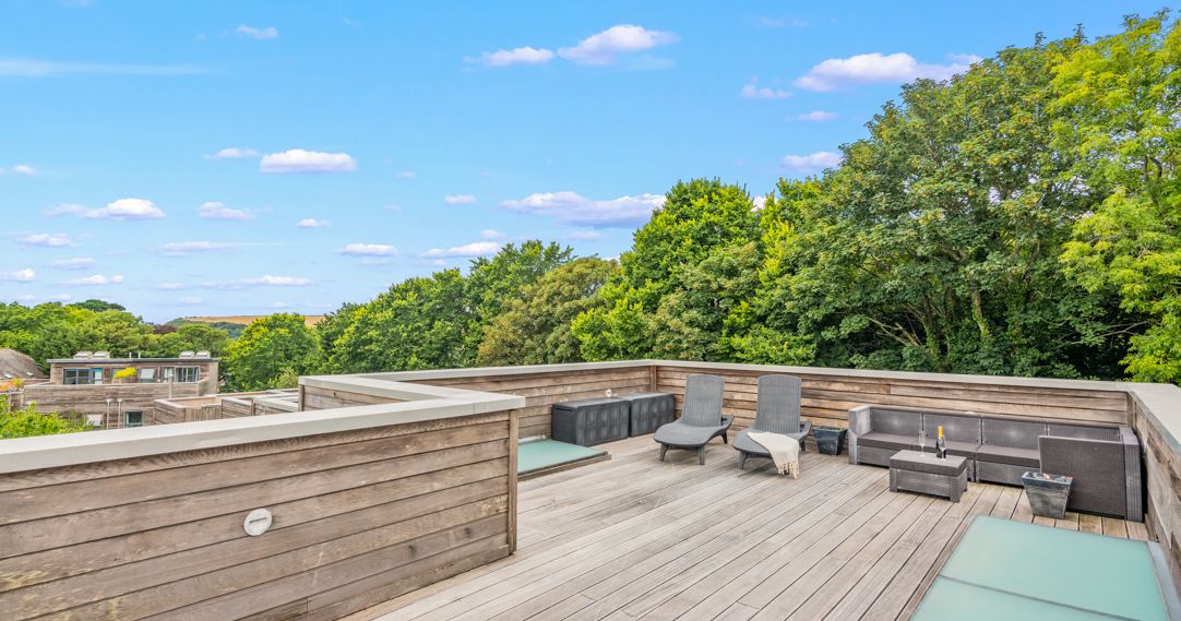 Spacious rooftop terrace with wooden decking, lounge chairs, a sectional sofa, and surrounding greenery under a blue sky with scattered clouds.