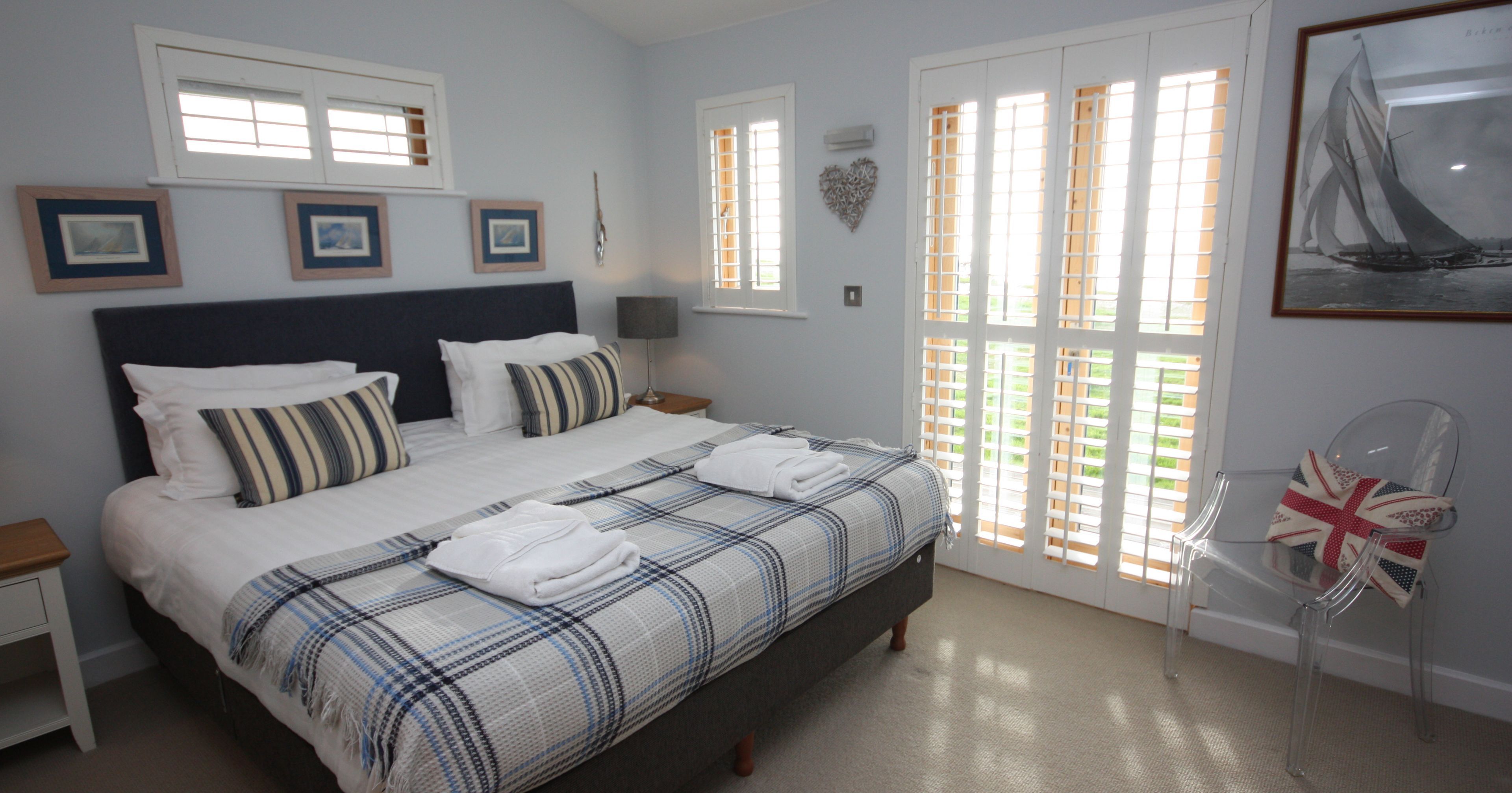 Bright bedroom with a double bed, striped pillows, and white linens, featuring large windows with wooden shutters.