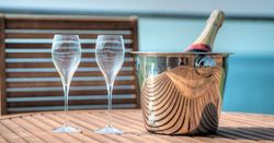 Two empty wine glasses and a bottle of champagne in a silver ice bucket on a wooden table outdoors.