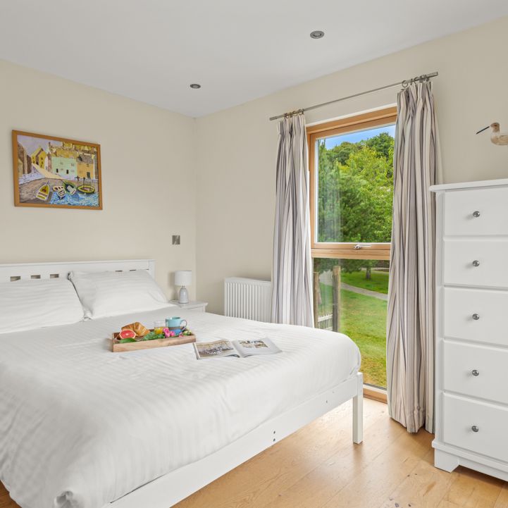 Bright and airy bedroom with a white bed, white furniture, wooden floor, a window with striped curtains showing a view of greenery, and a breakfast tray on the bed.