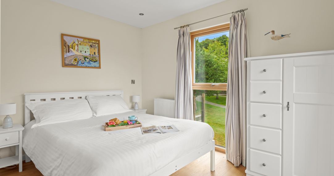 Bright and airy bedroom with a white bed, white furniture, wooden floor, a window with striped curtains showing a view of greenery, and a breakfast tray on the bed.
