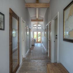 Long modern hallway with stone tile floor, wooden ceiling beams, wall art and a large mirror, leading to a sunlit sitting area.