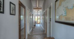 Long modern hallway with stone tile floor, wooden ceiling beams, wall art and a large mirror, leading to a sunlit sitting area.