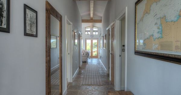 Long modern hallway with stone tile floor, wooden ceiling beams, wall art and a large mirror, leading to a sunlit sitting area.
