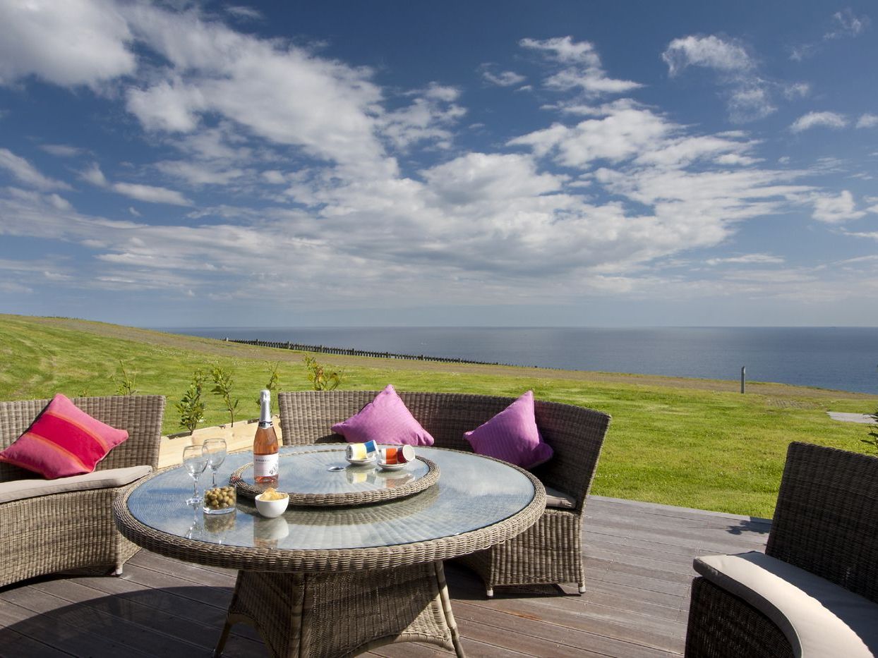Outdoor wicker patio set with a glass table and colorful pillows, overlooking a grassy field and the ocean under a blue sky.