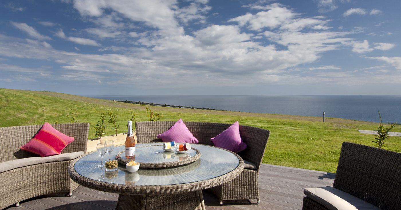 Outdoor wicker patio set with a glass table and colorful pillows, overlooking a grassy field and the ocean under a blue sky.