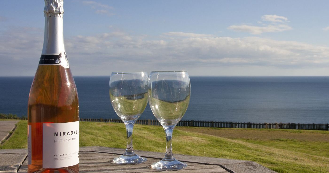 A bottle of Mirabello pinot grigio rosé and two wine glasses on a wooden table with an ocean view in the background.