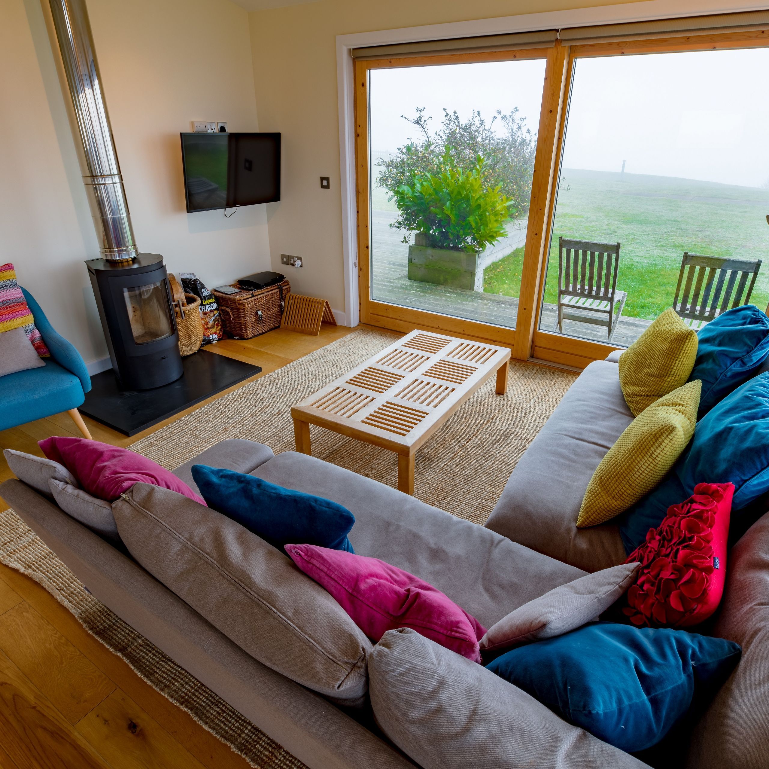 Modern cozy living room with colorful cushions, a wood-burning stove, and large windows overlooking green grass and patio chairs.