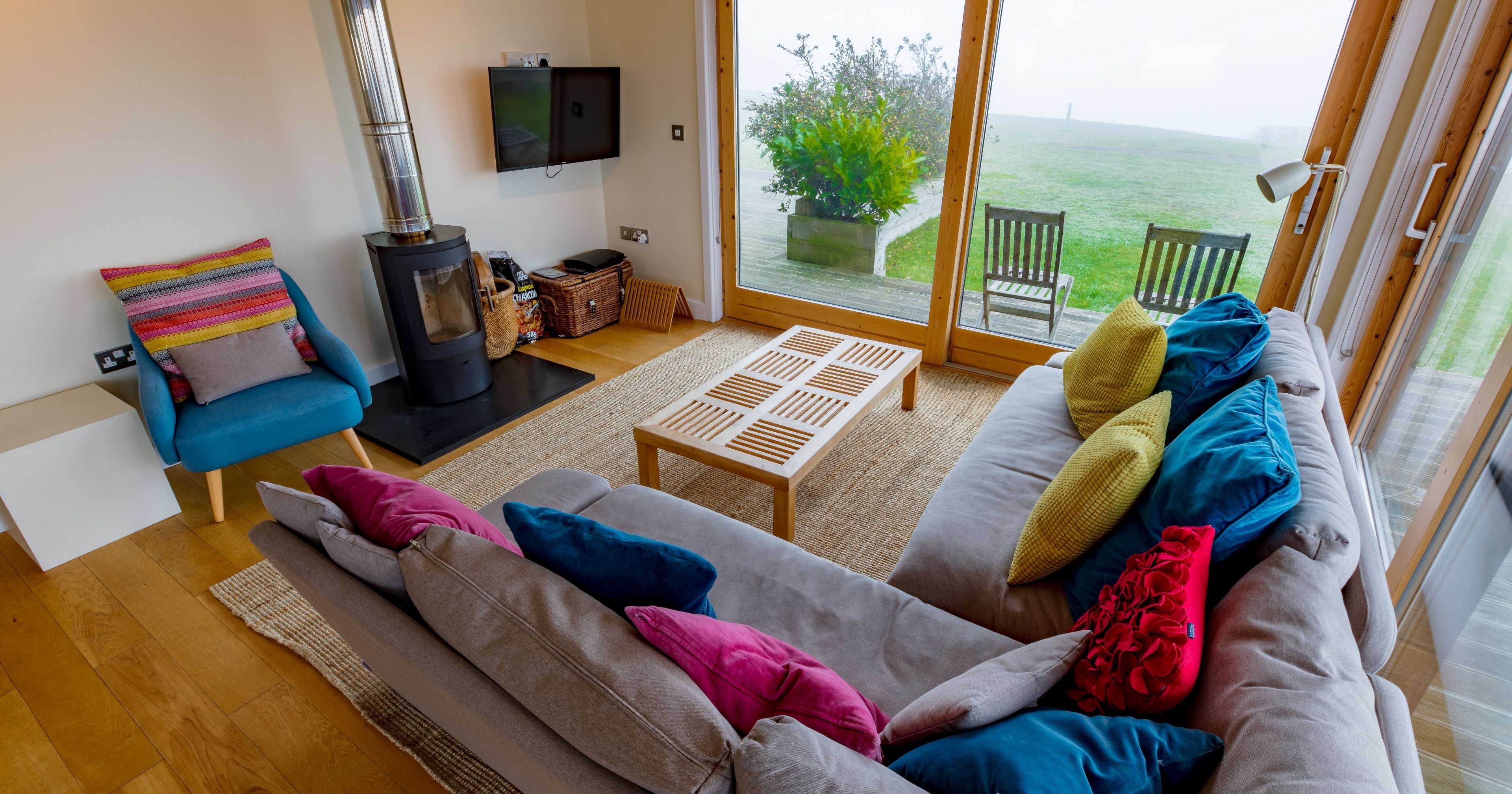 Modern cozy living room with colorful cushions, a wood-burning stove, and large windows overlooking green grass and patio chairs.