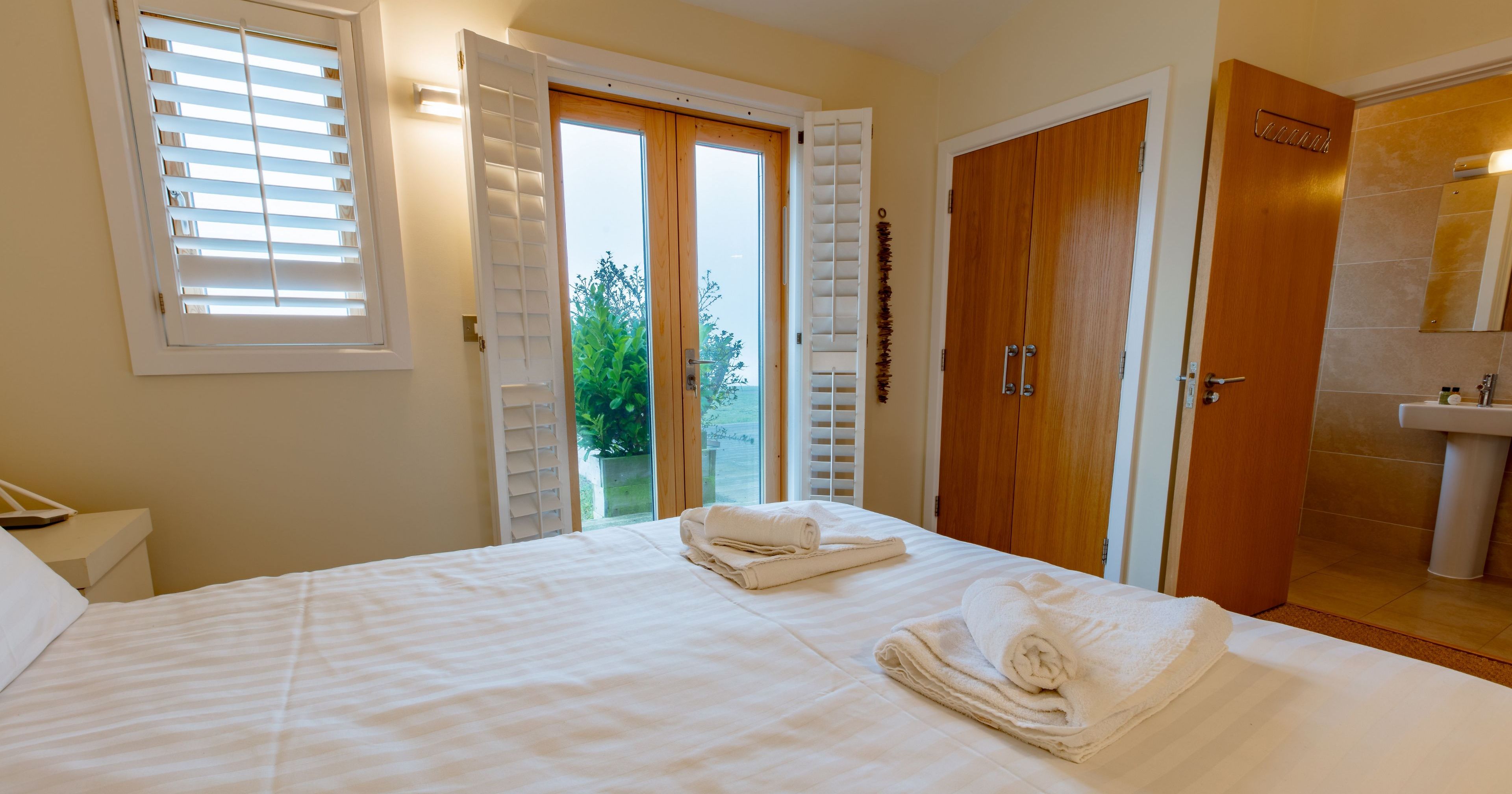 Neatly made bed in a cozy bedroom with white linens and folded towels, next to patio doors and an open bathroom door.