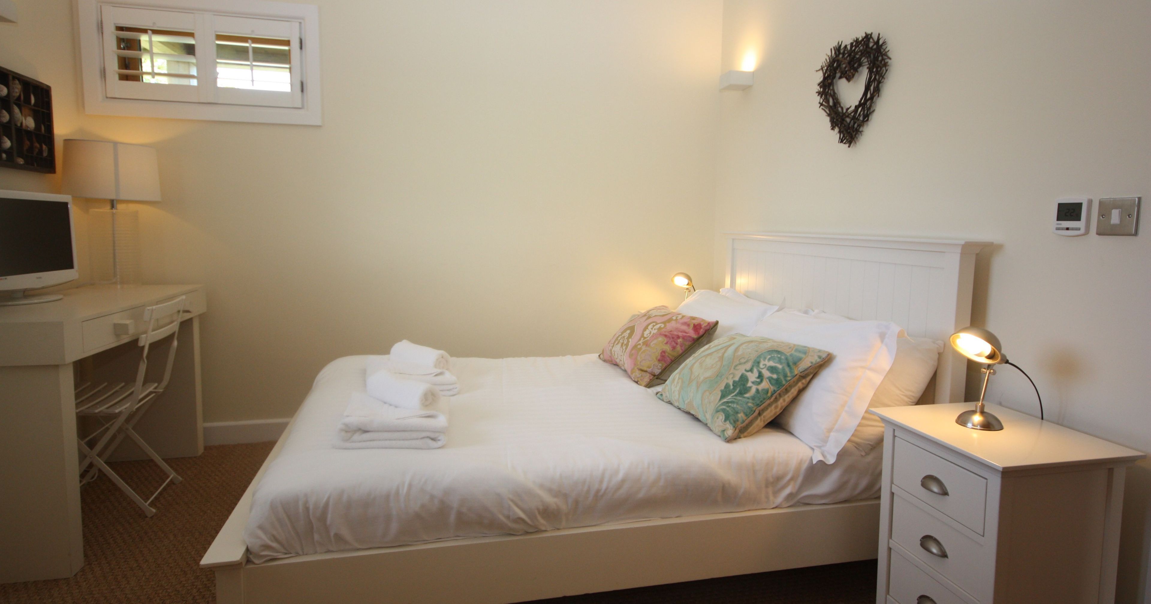Bright, modern bedroom with a white bed, colorful pillows, and a small desk with a lamp and TV.