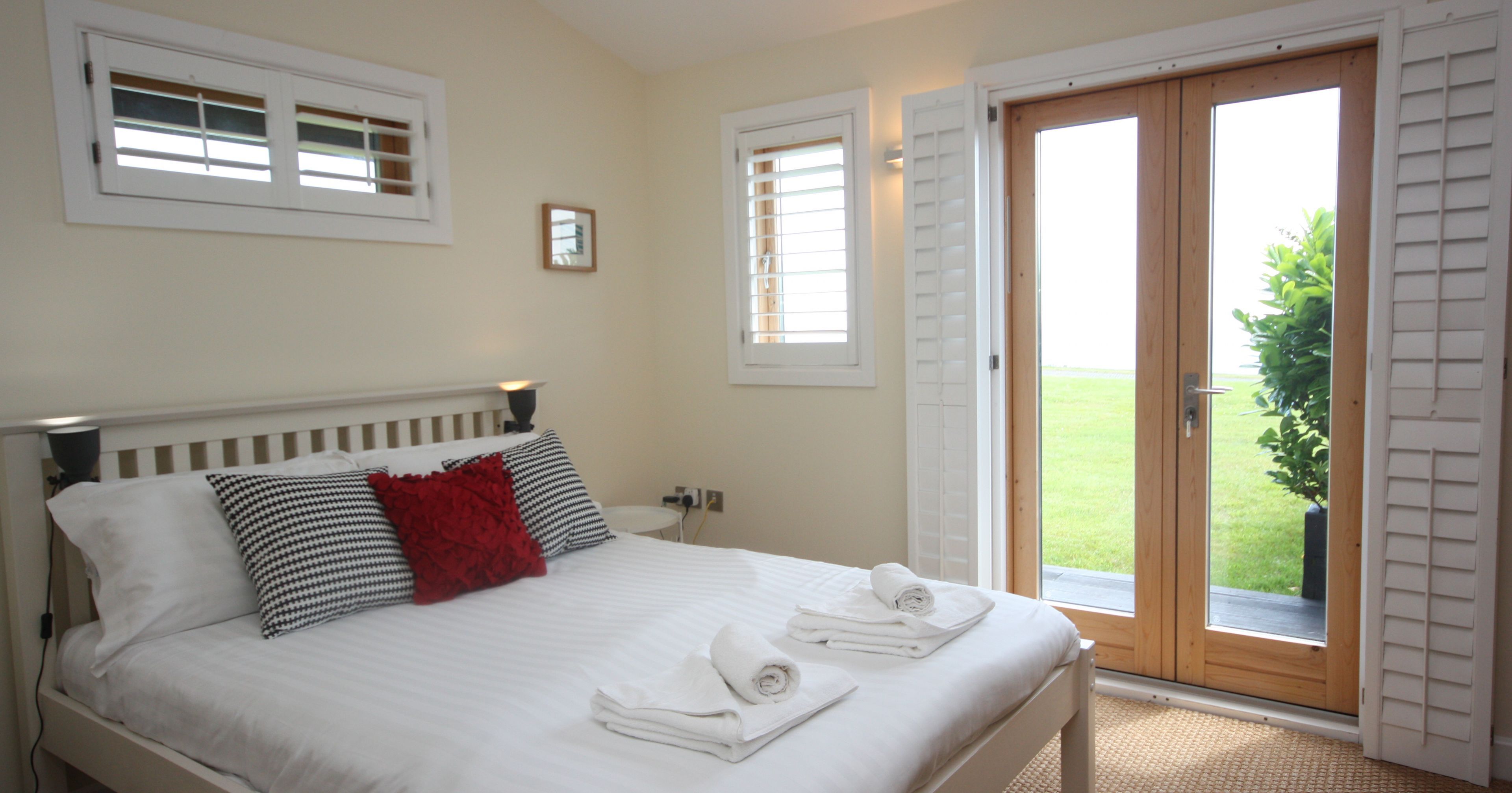 Bright bedroom with white bed linens, accent pillows, and French doors opening to a green lawn