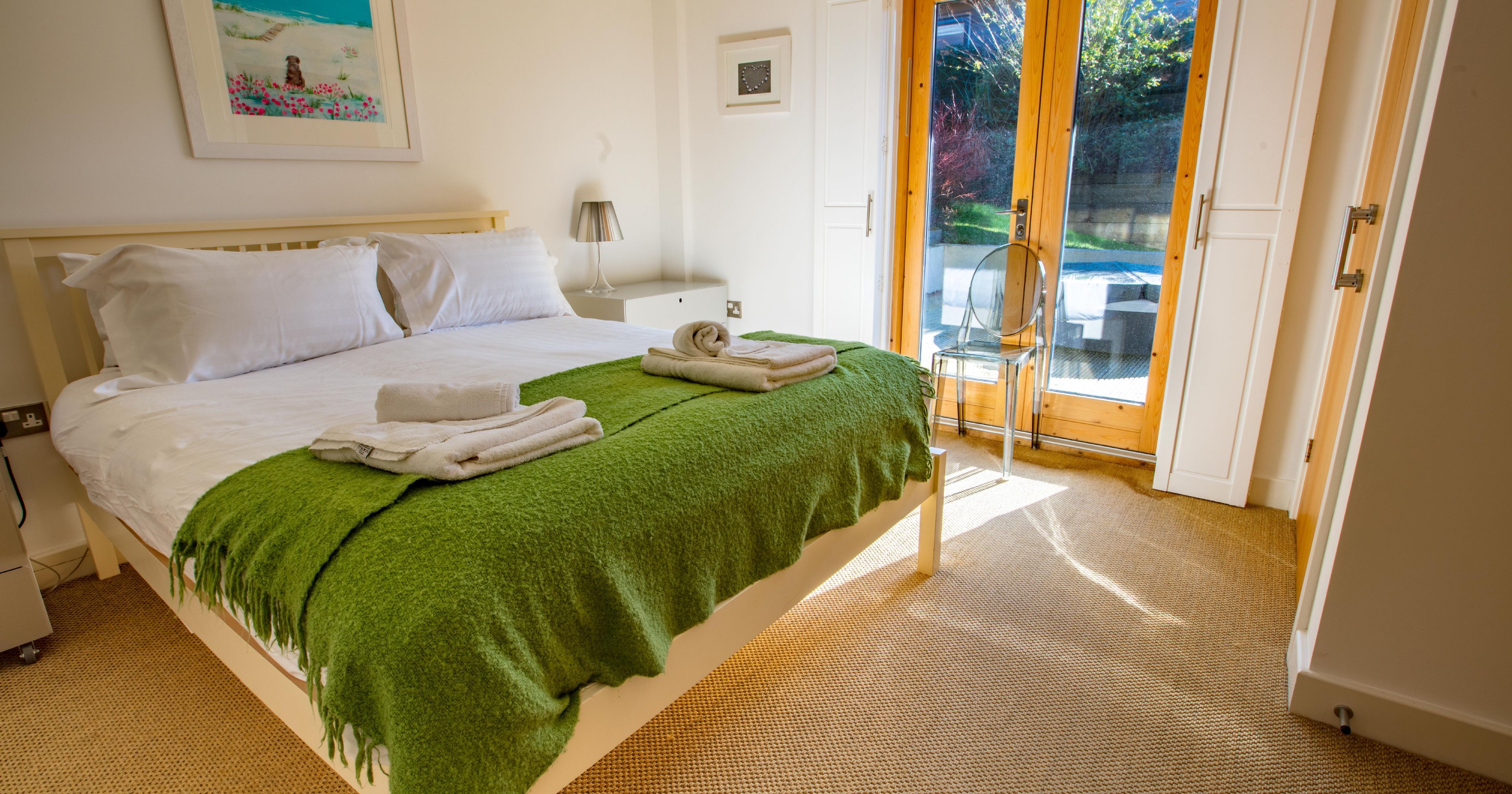 Bright modern bedroom with a double bed, green throw blanket, and folded towels by glass doors leading outside.