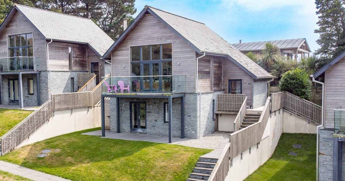 Modern two-story house with wooden and stone exterior, balcony, and sloped landscaped yard.