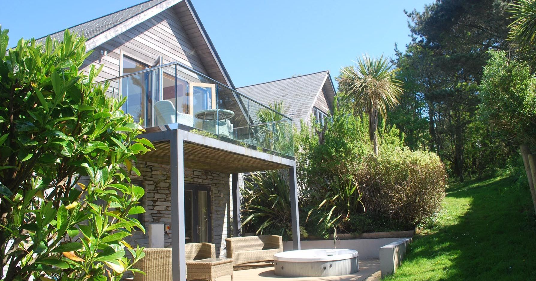 Modern house with upper balcony, outdoor seating, and hot tub surrounded by garden greenery