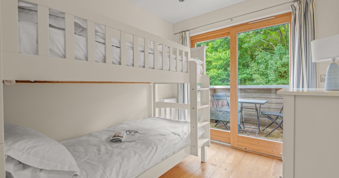 A bright bedroom with white bunk beds, wooden floor, a dresser with a lamp, and large glass doors opening to a balcony with a table and chairs overlooking green trees.