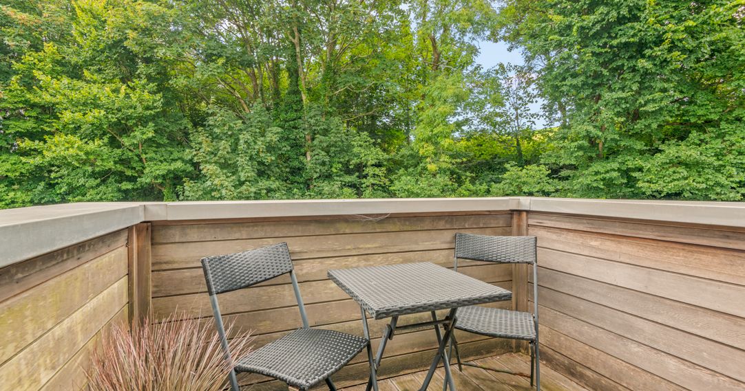 Small outdoor patio with two black wicker chairs and a square table, surrounded by wooden fencing and overlooking lush green trees.