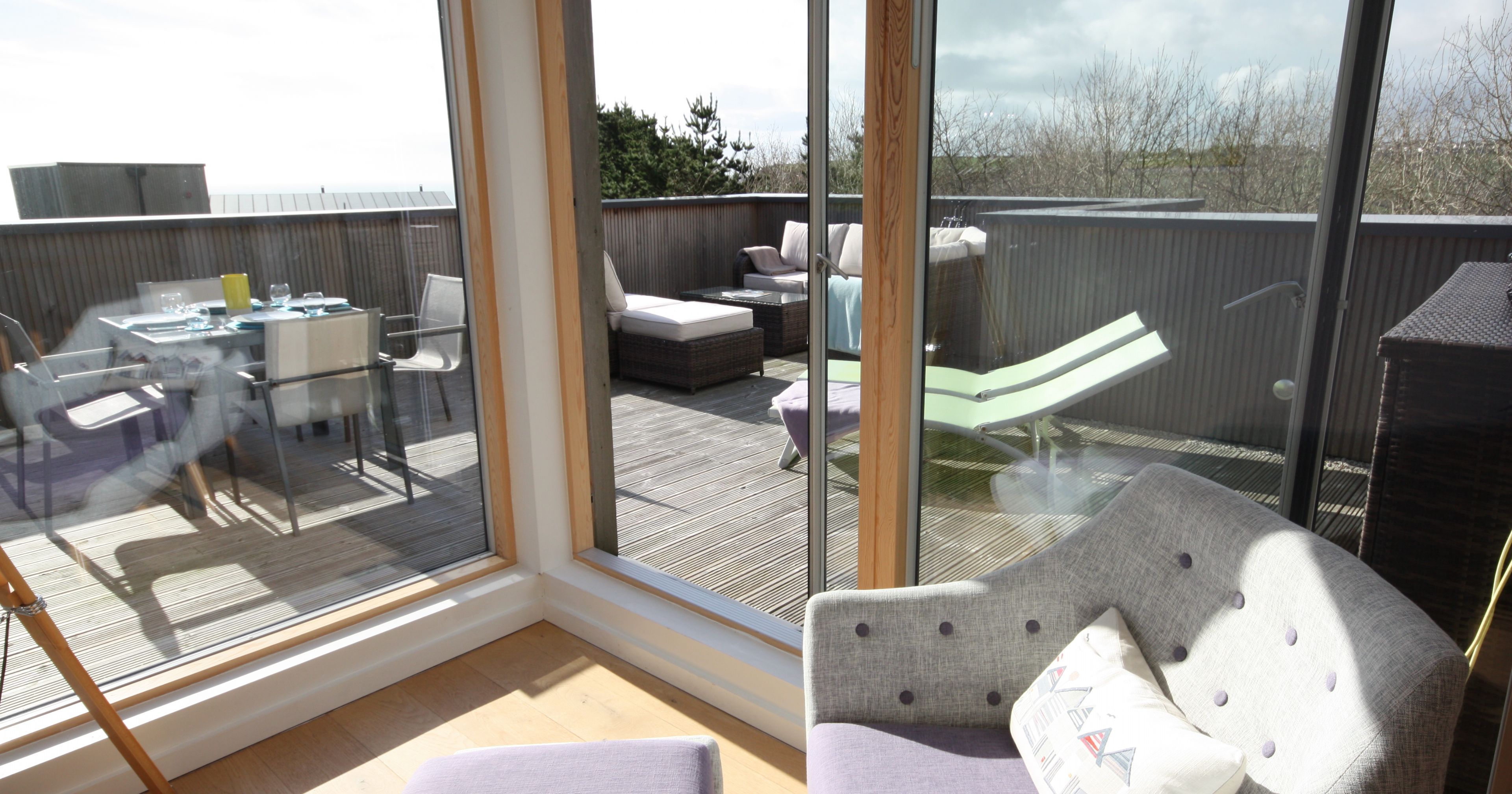 View through large windows of a sunny patio with outdoor furniture including chairs, a table, and loungers.