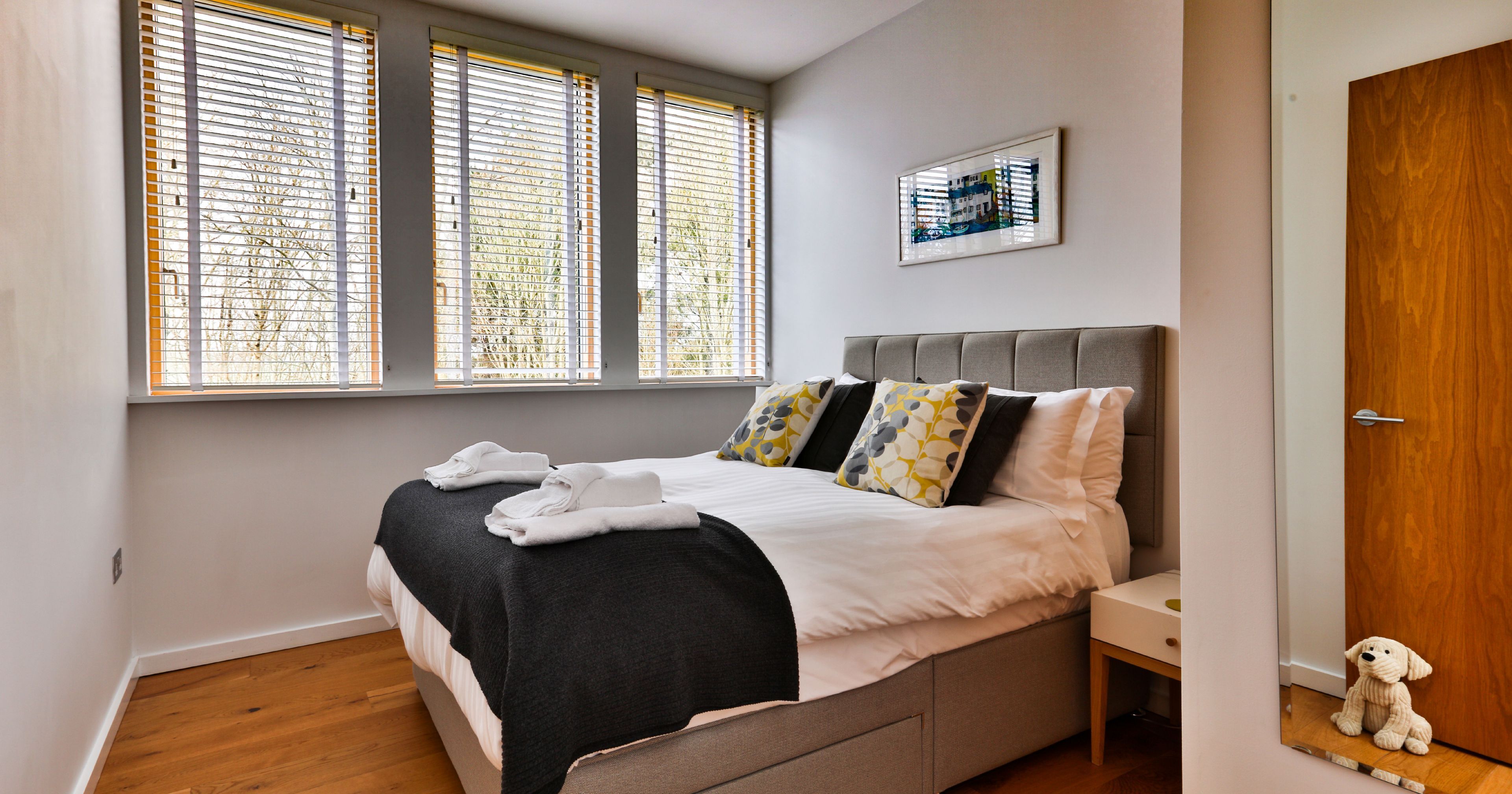 Modern bedroom with a large bed, decorative pillows, and three windows with blinds.