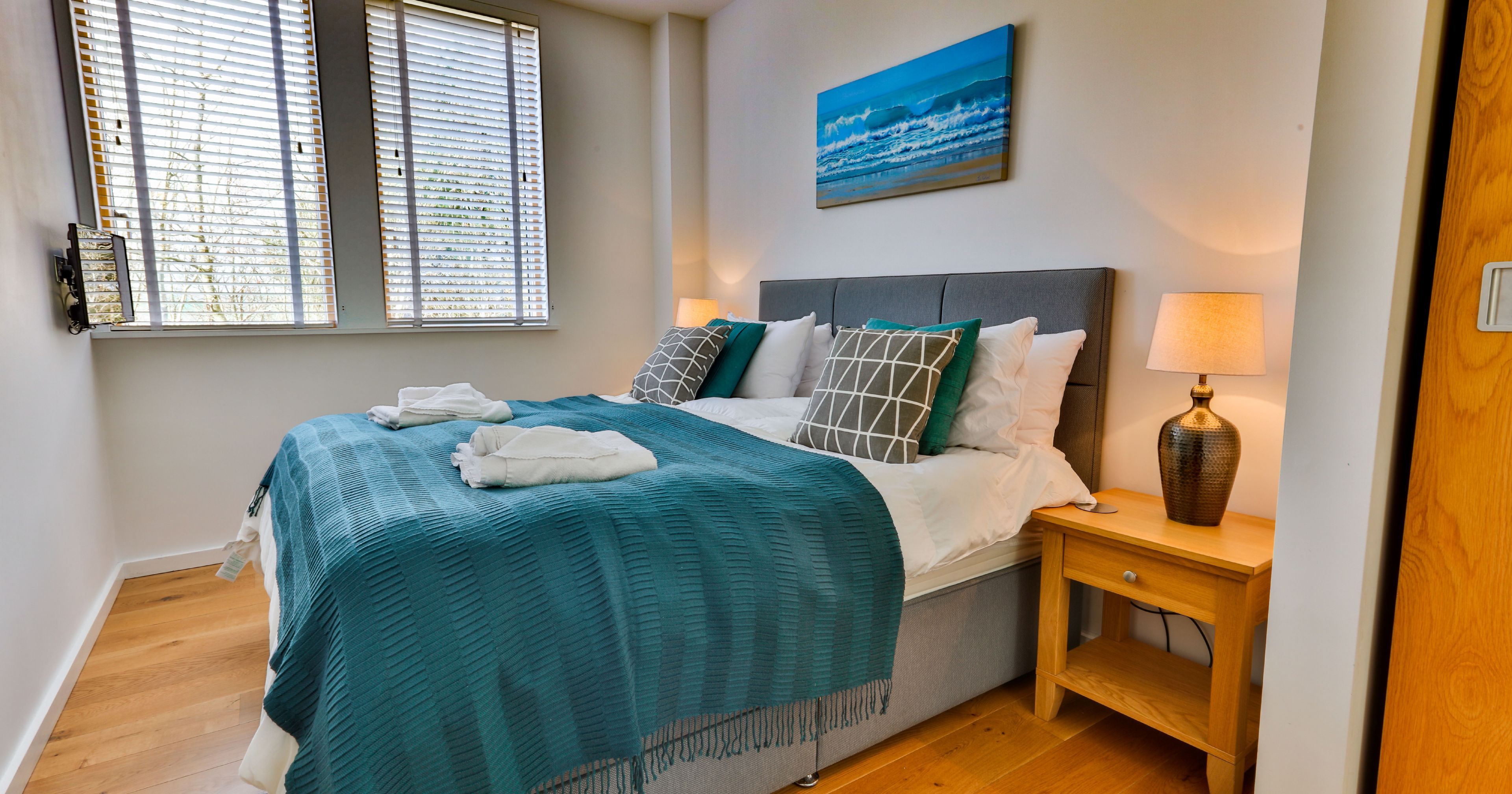 Modern bedroom with a teal blanket, large windows, and bedside lamp