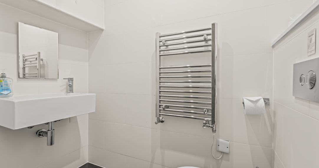 Modern bathroom with white walls, a wall-mounted sink, mirror, chrome towel radiator, toilet, and toilet paper holder.