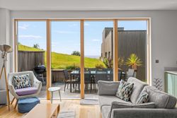 Modern living room with large glass doors opening to a patio with outdoor dining area and grassy hill view