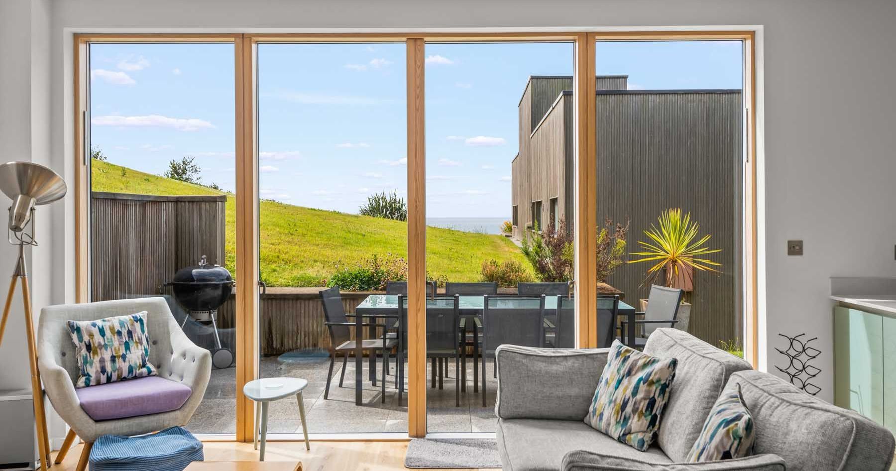 Modern living room with large glass doors opening to a patio with outdoor dining area and grassy hill view