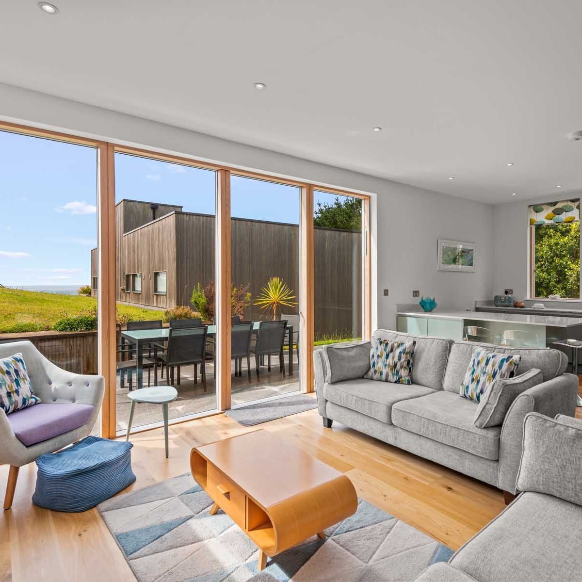 Modern open plan living room and kitchen with large windows and patio doors overlooking a green garden.