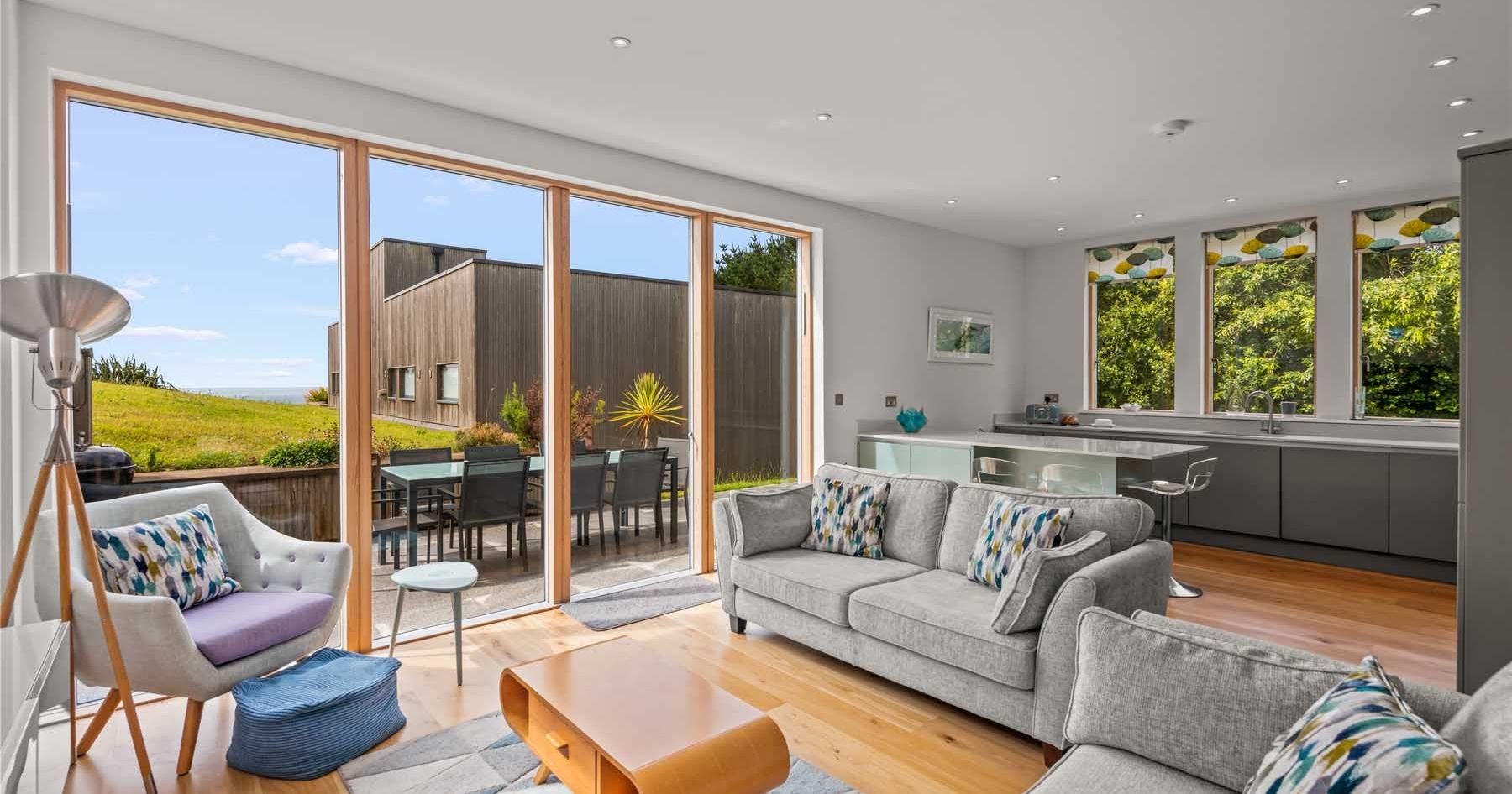 Modern open plan living room and kitchen with large windows and patio doors overlooking a green garden.