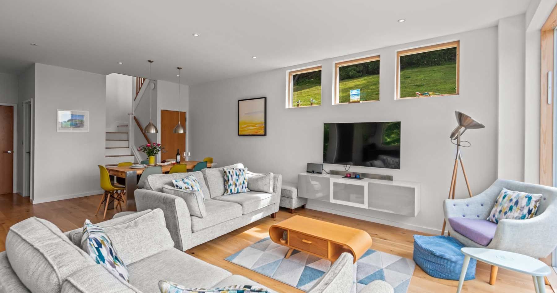 Modern living room and dining area with light grey sofas, flat screen TV, and large windows.