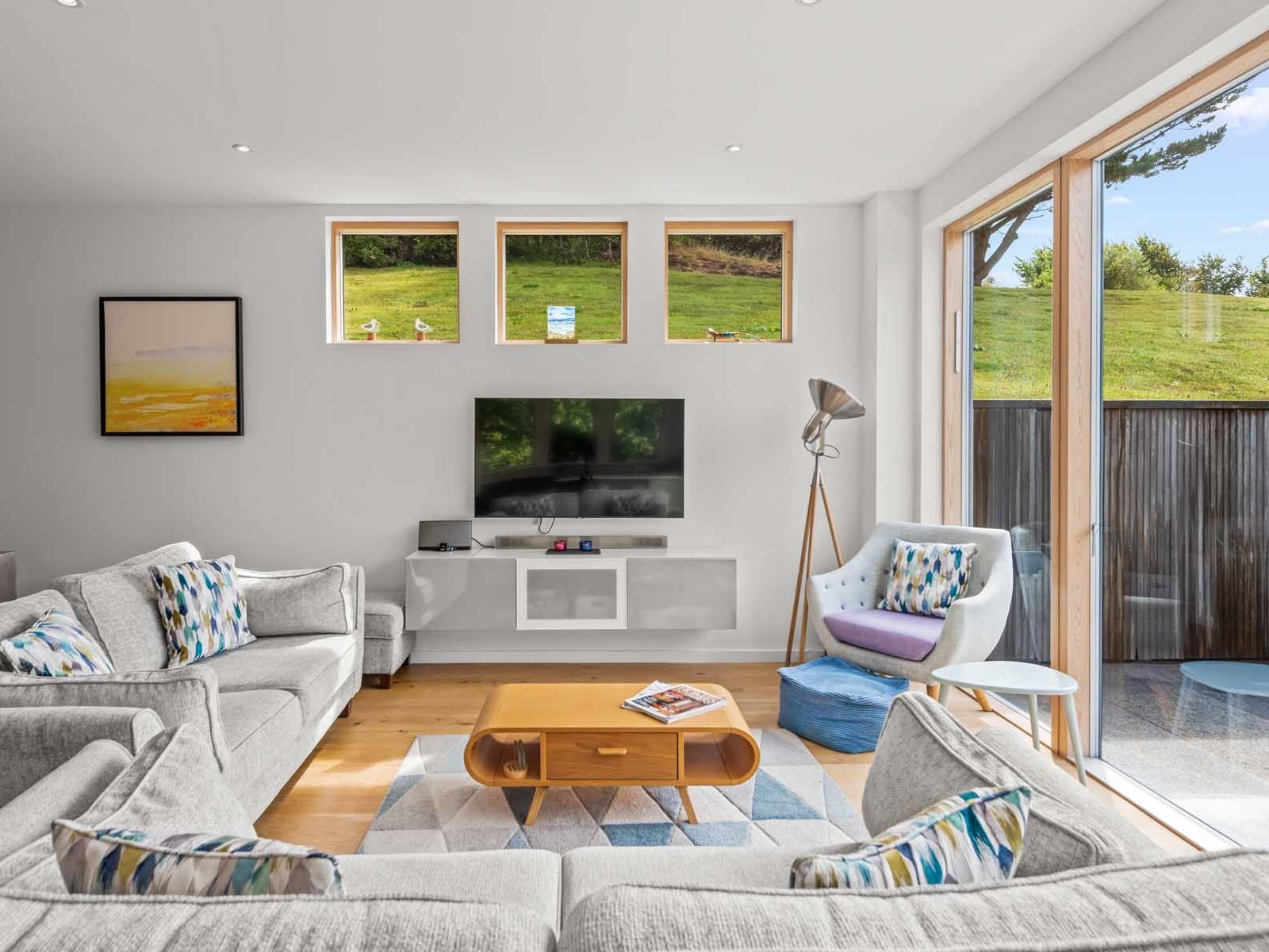 Modern living room with large windows, light grey furniture, and a wall-mounted TV.