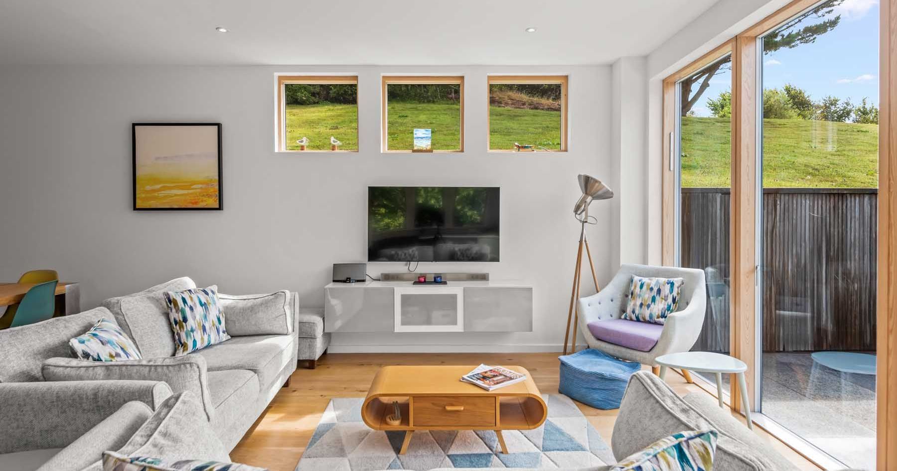 Modern living room with large windows, light grey furniture, and a wall-mounted TV.