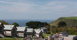 Modern wooden houses with sloped roofs overlooking the ocean and green hillside under a partly cloudy sky.