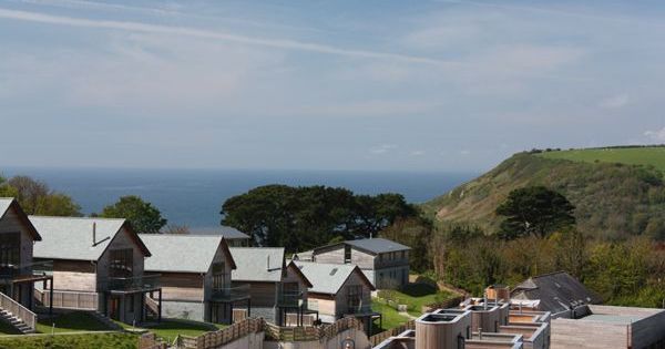 Modern wooden houses with sloped roofs overlooking the ocean and green hillside under a partly cloudy sky.