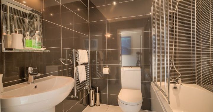 Modern bathroom with dark glossy tiles, white sink, toilet, bathtub with shower, and wall-mounted mirror.