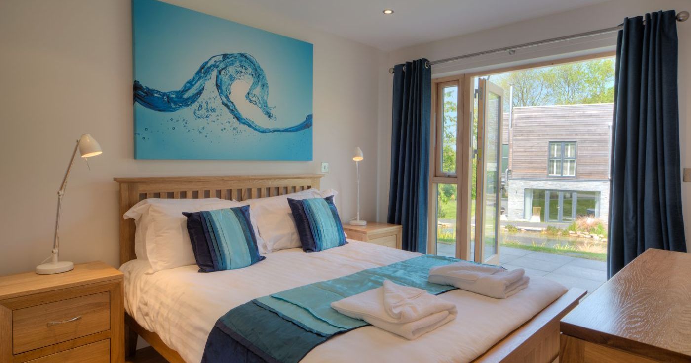 Modern bedroom with wooden furniture, blue and white bedding, abstract water-themed artwork above the bed, and patio doors opening to a garden view.