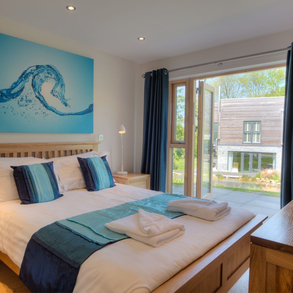 Modern bedroom with wooden furniture, blue and white bedding, abstract water-themed artwork above the bed, and patio doors opening to a garden view.