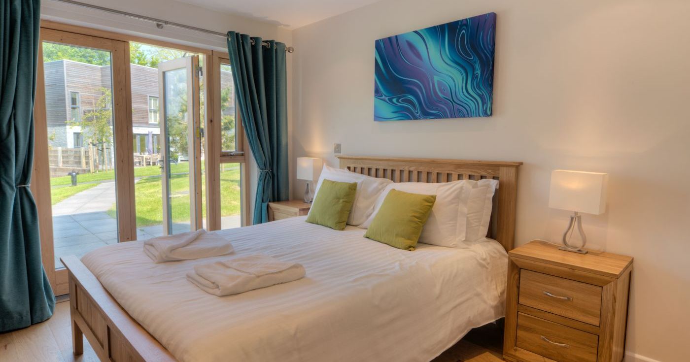 Modern bedroom with a double bed, green pillows, wooden furniture, and large windows overlooking a garden.