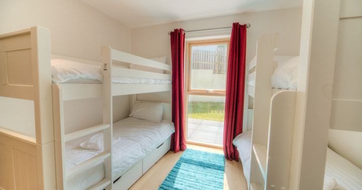 Bright bedroom with two sets of white bunk beds, a window with red curtains, and a blue rug on the floor.