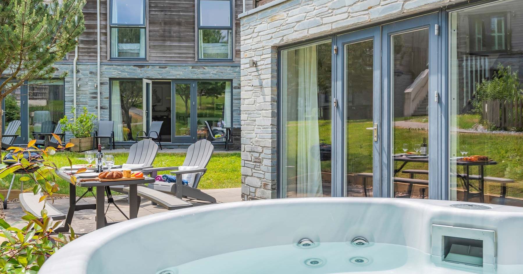 Outdoor patio with a hot tub, dining table, and wooden chairs in front of a modern house.