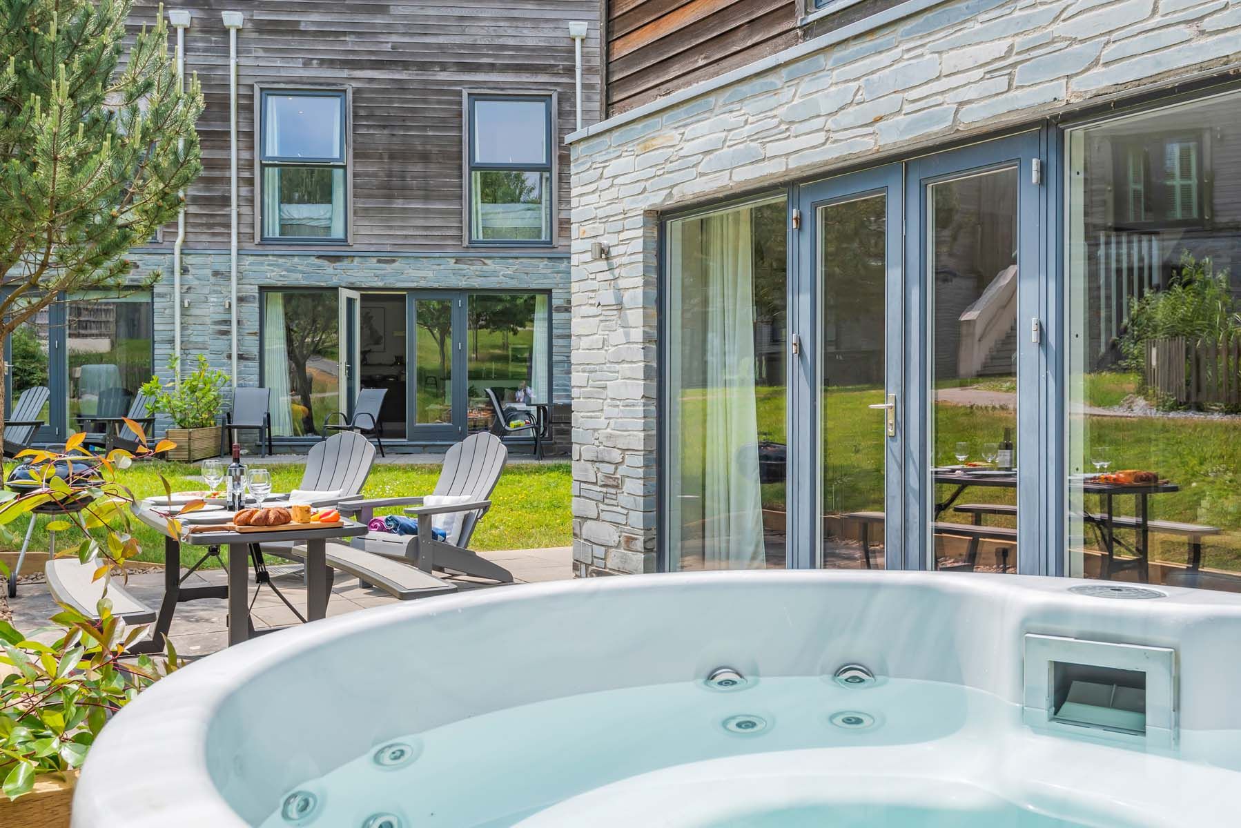Outdoor patio with a hot tub, dining table, and wooden chairs in front of a modern house.