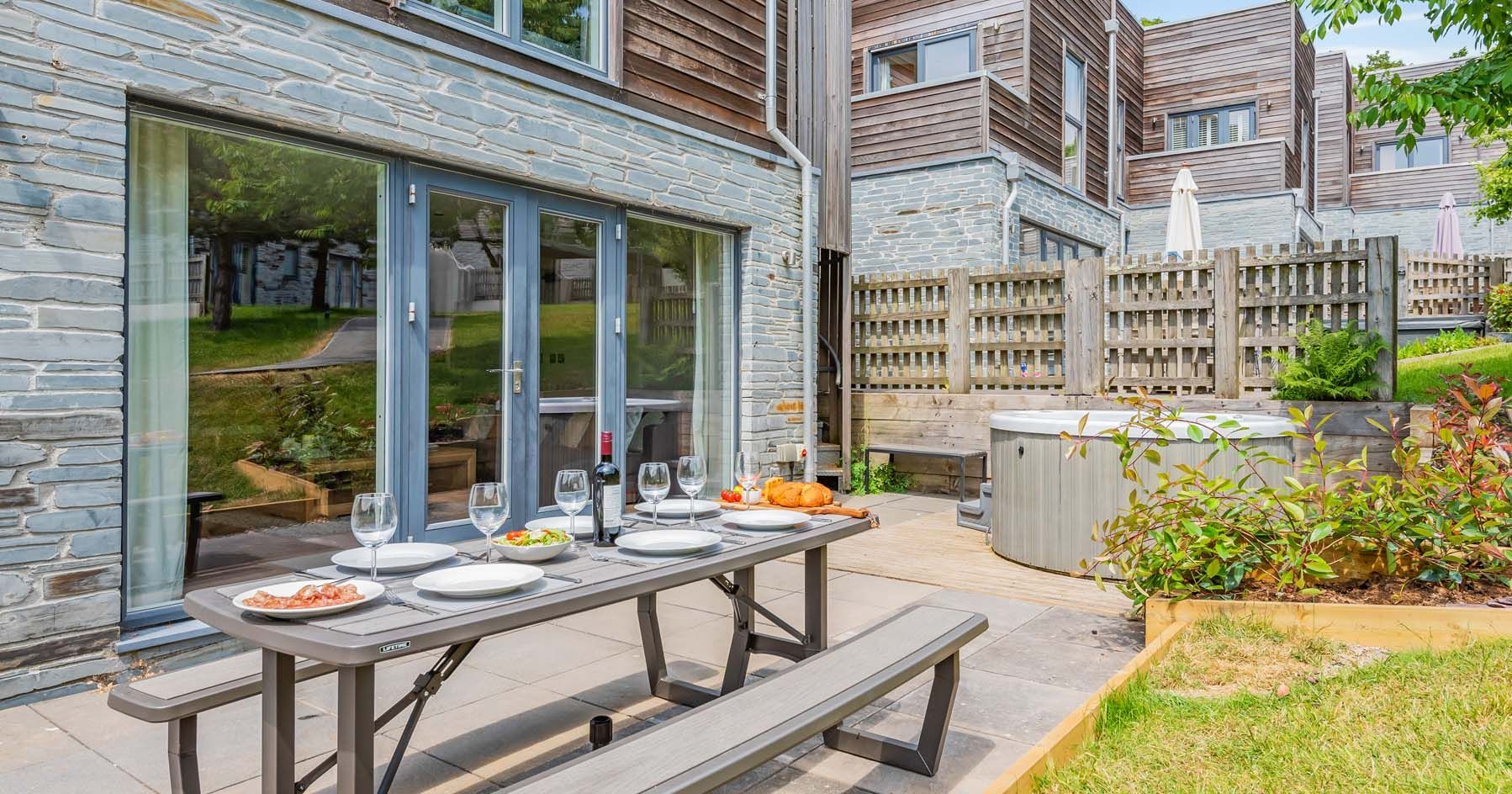 Modern outdoor patio with picnic table, set for four, next to a hot tub and wooden-fenced yard.