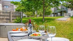 Outdoor dining table set with plates, wine glasses, a bottle of wine, salad, and bread in a garden with modern townhouses in the background.