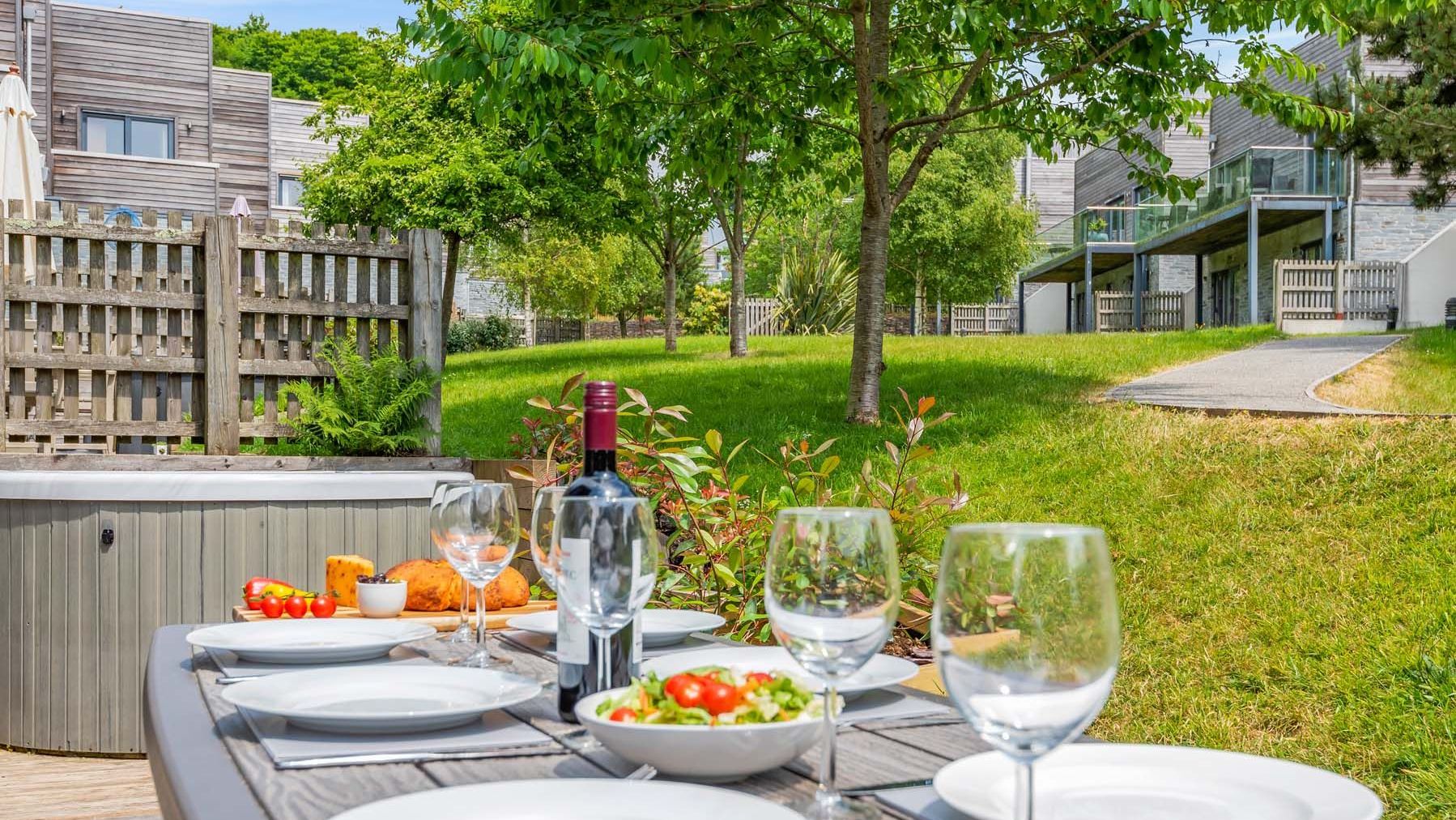 Outdoor dining table set with plates, wine glasses, a bottle of wine, salad, and bread in a garden with modern townhouses in the background.