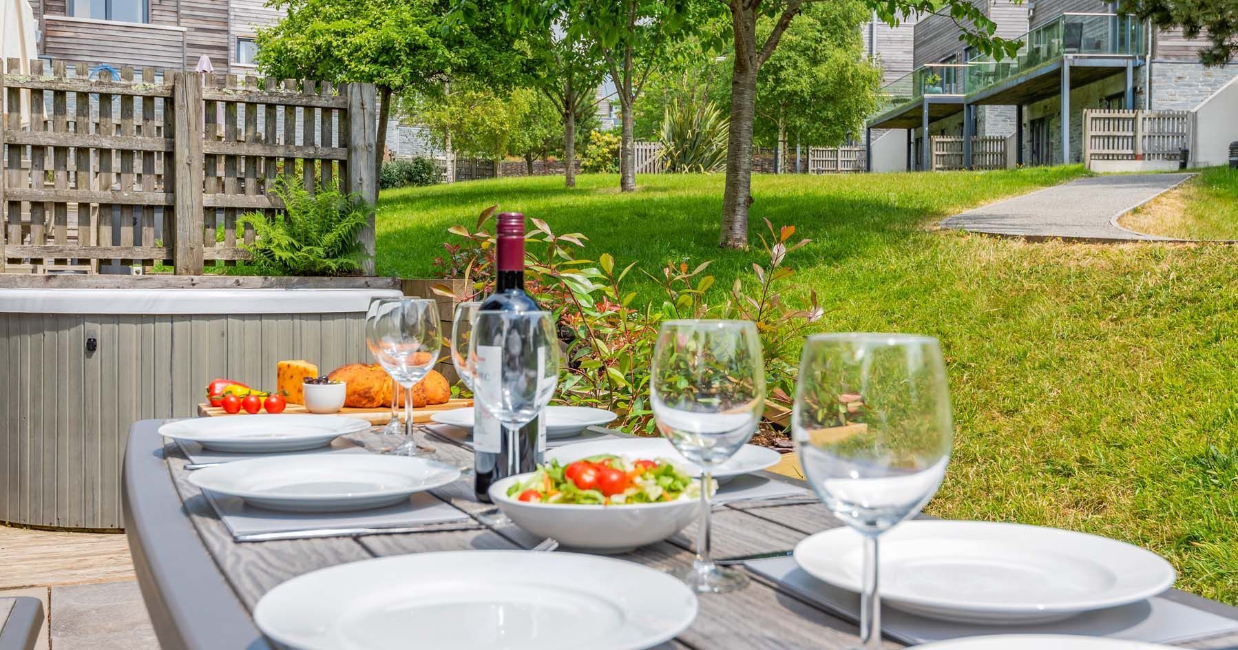 Outdoor dining table set with plates, wine glasses, a bottle of wine, salad, and bread in a garden with modern townhouses in the background.