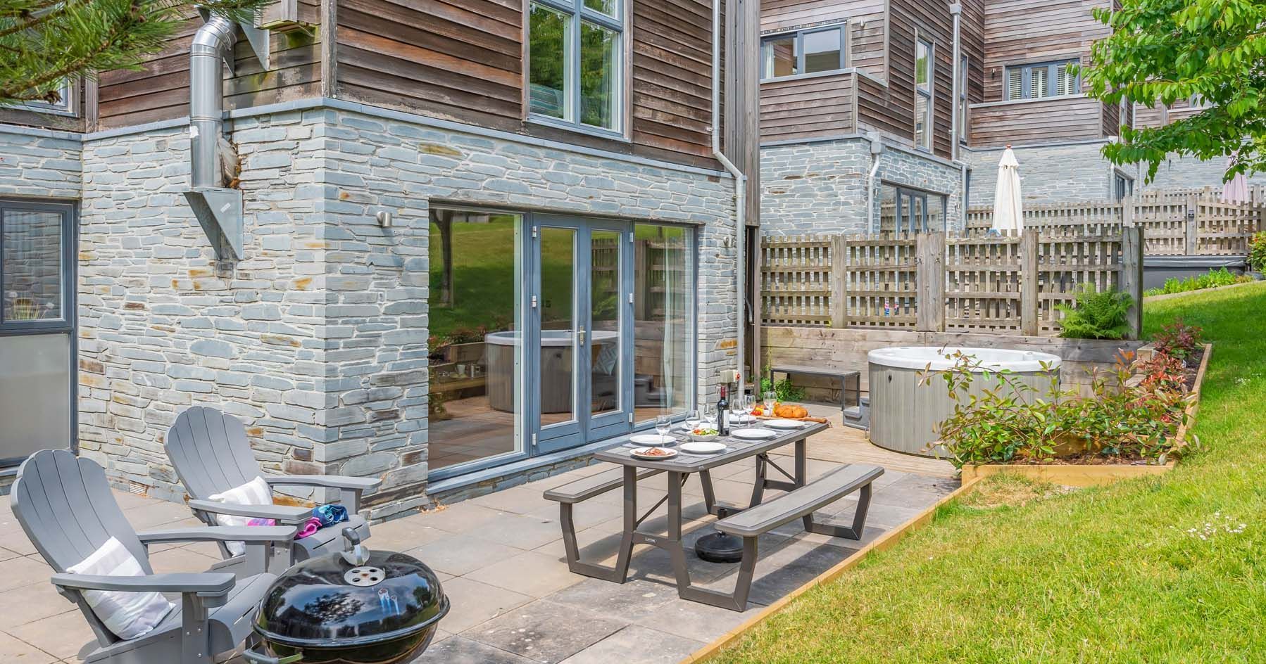 Modern patio area with outdoor seating, dining table, barbecue grill, and hot tub outside a stone and wood house.