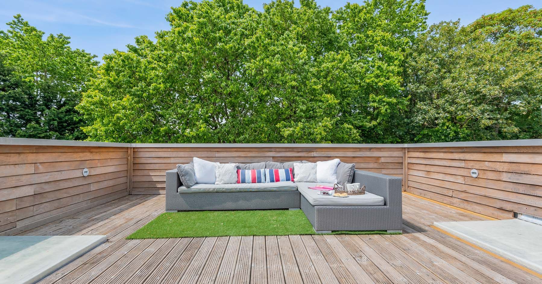 Modern rooftop terrace with wooden decking, a grey outdoor sectional sofa, and green trees in the background.