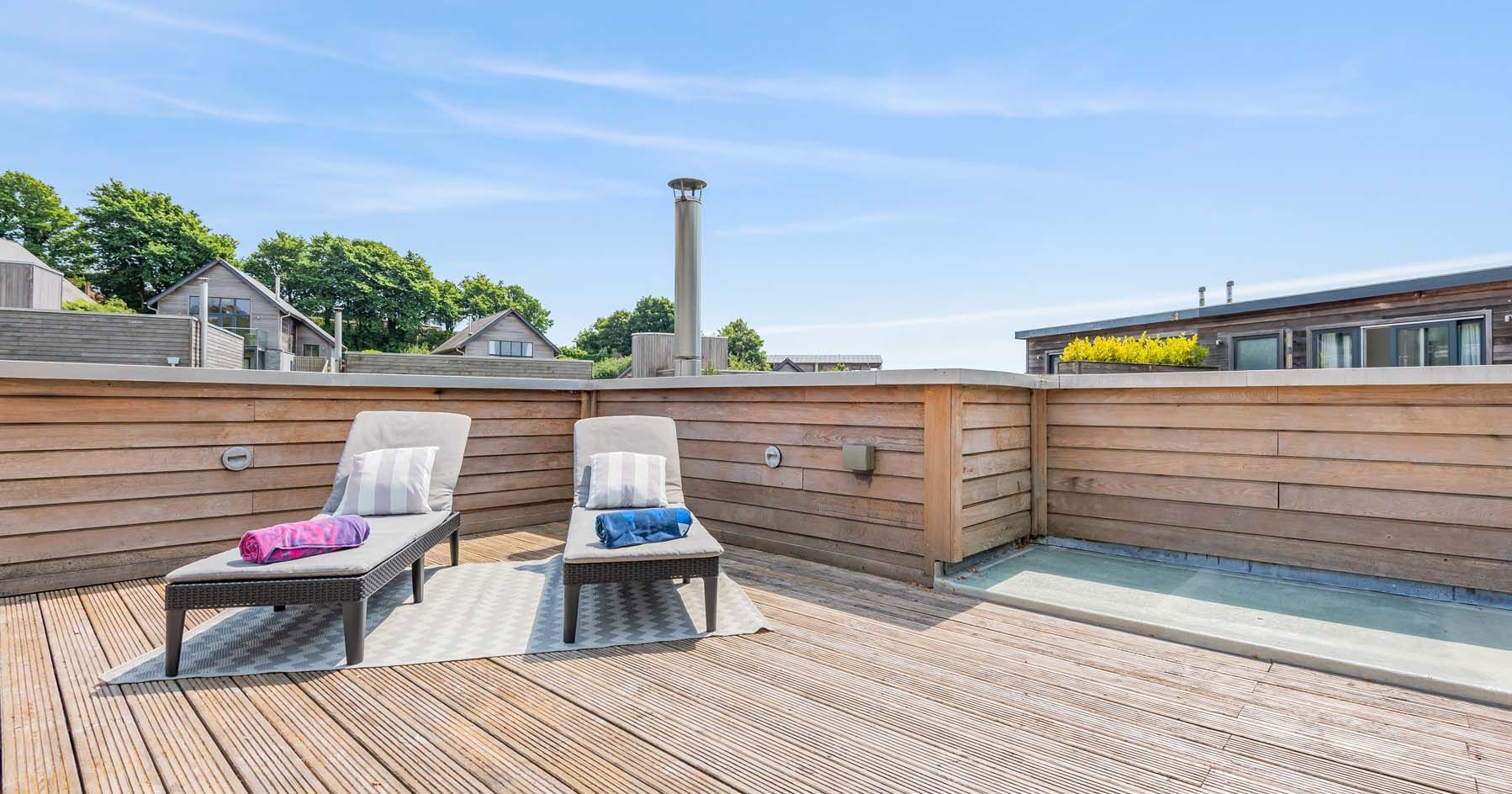 Two lounge chairs on a wooden rooftop deck with towels and pillows, surrounded by a wooden fence.